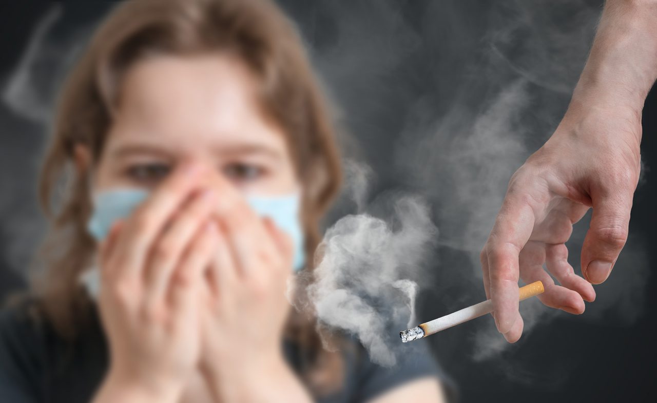 Dziewczynka w maseczce zasłania nos i usta. Obok dłoń trzymająca zapalonego papierosa.